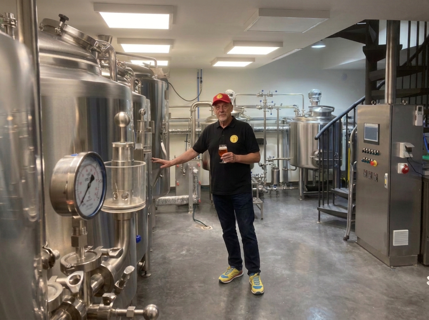 Brouwerijruimte met man in rode pet die bier vasthoudt naast roestvrijstalen tanks en leidingen.