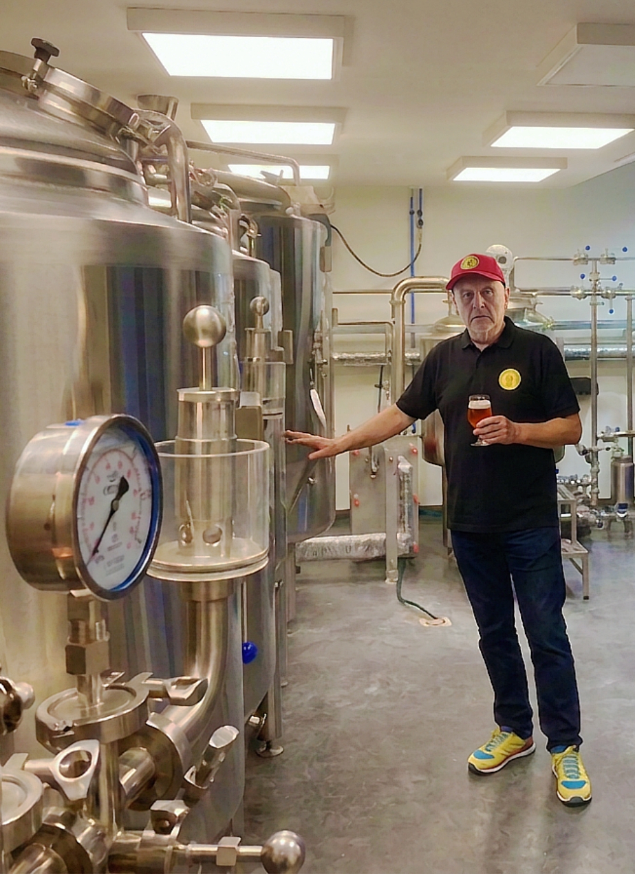 Brouwerijruimte met man in rode pet die bier vasthoudt naast roestvrijstalen tanks en leidingen.